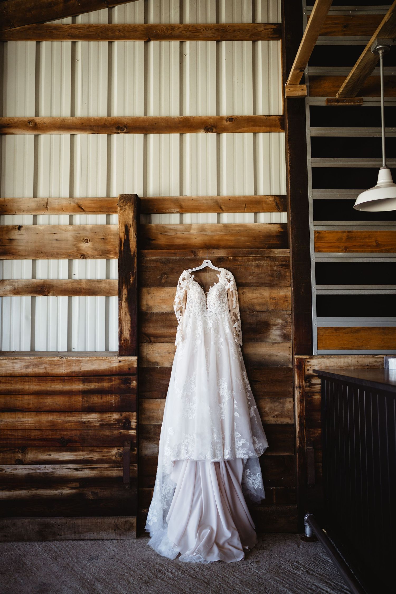A beautiful lace wedding dress with long sleeves hangs on rustic wooden barn wall next to black stairs.