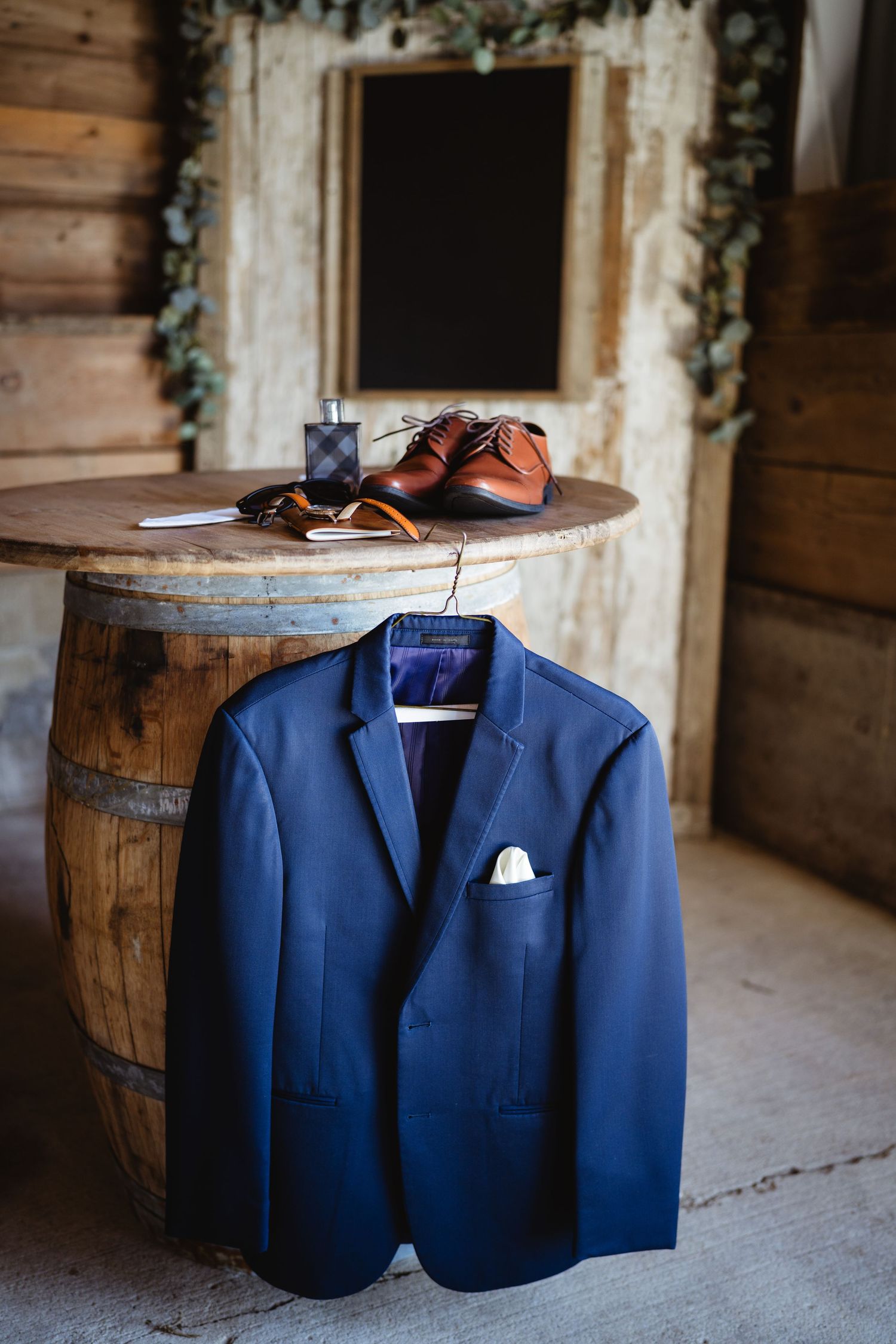 A navy blue groom's suit jacket hangs near a rustic wooden barrel topped with brown leather dress shoes and accessories.