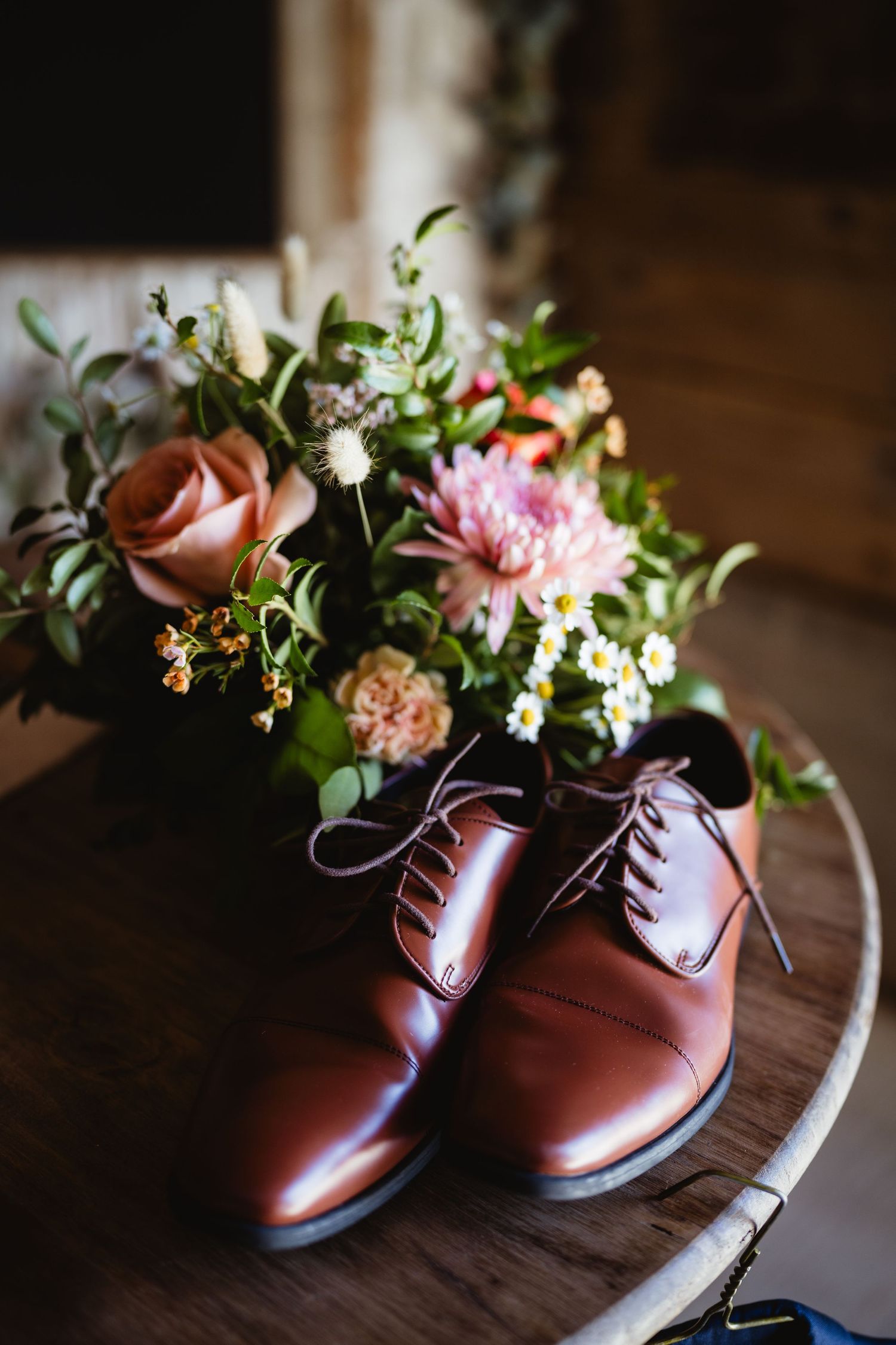 Brown leather dress shoes and a fresh floral arrangement with pink roses sit on a dark wooden surface.