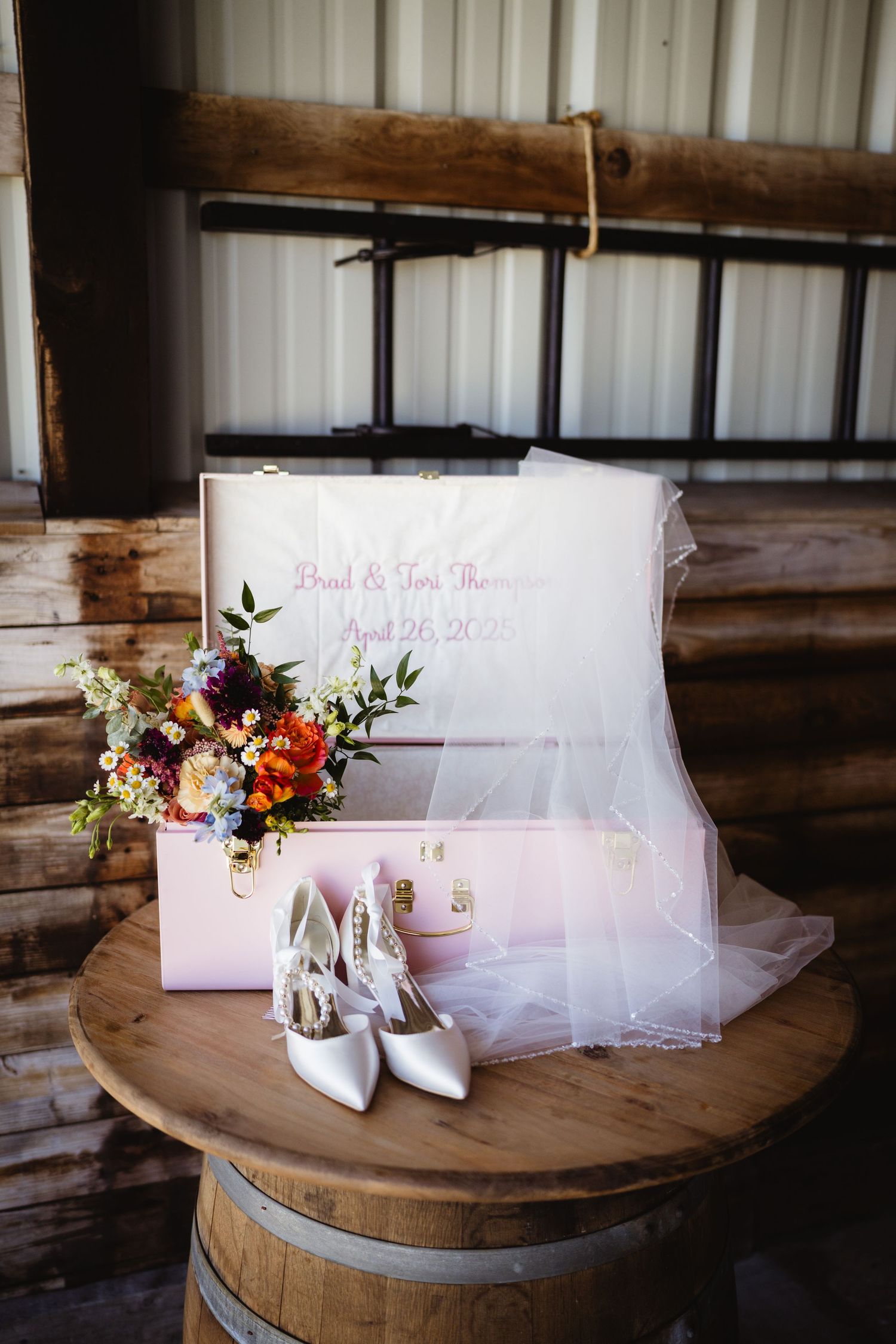 White bridal heels and wedding box with delicate tulle veil sit atop rustic wooden barrel in barn setting.
