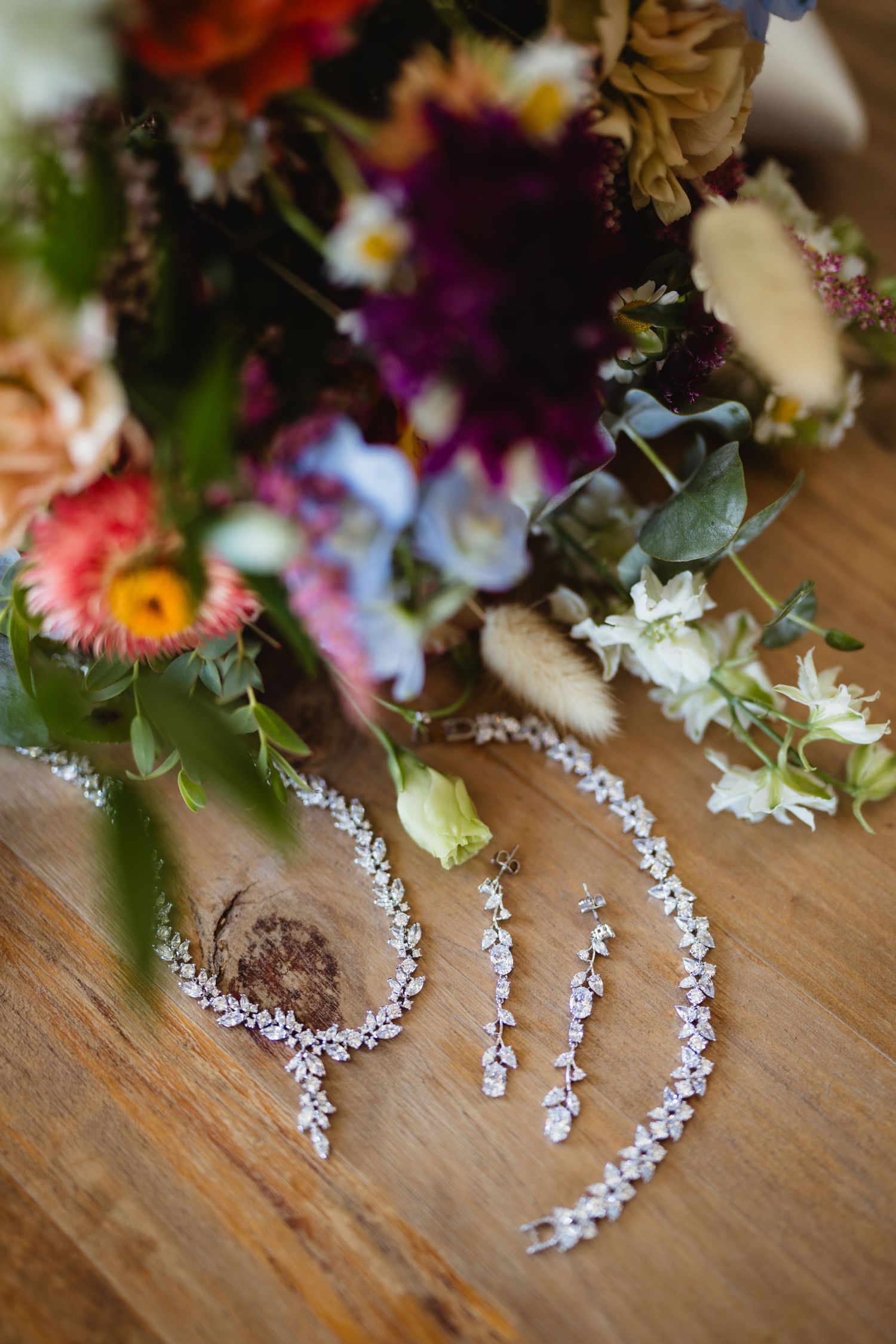 Elegant crystal bridal jewelry and accessories laid out on wooden surface next to colorful wedding flower bouquet.
