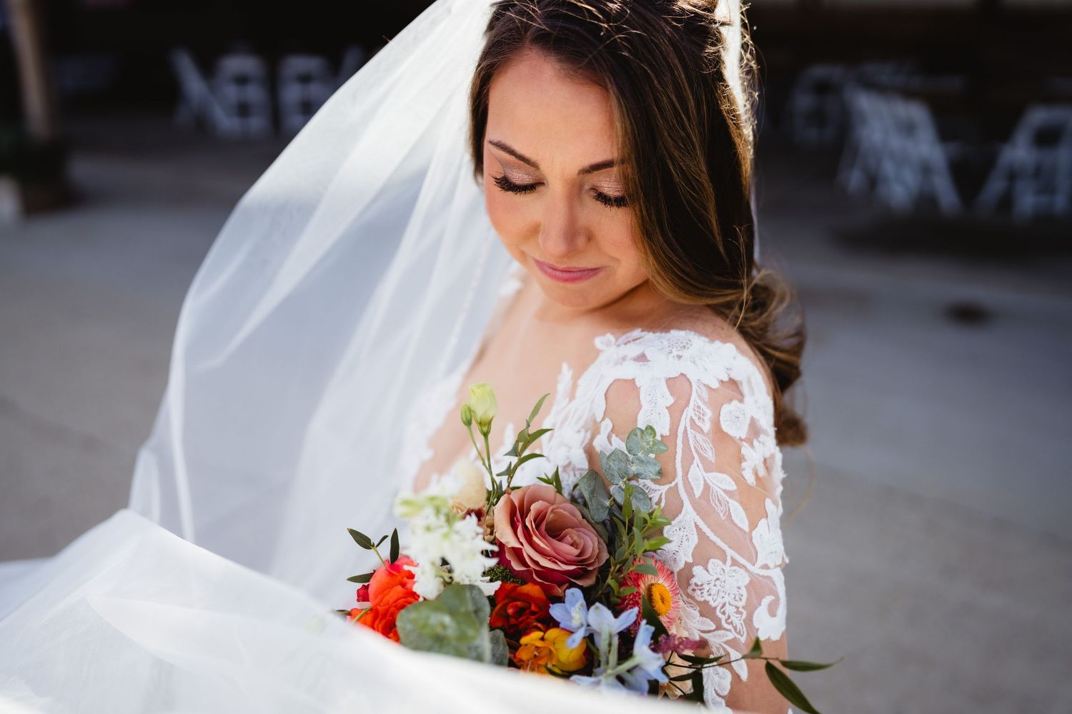 Beautiful bride in lace dress looking down at colorful bouquet of roses and flowers on her wedding day.
