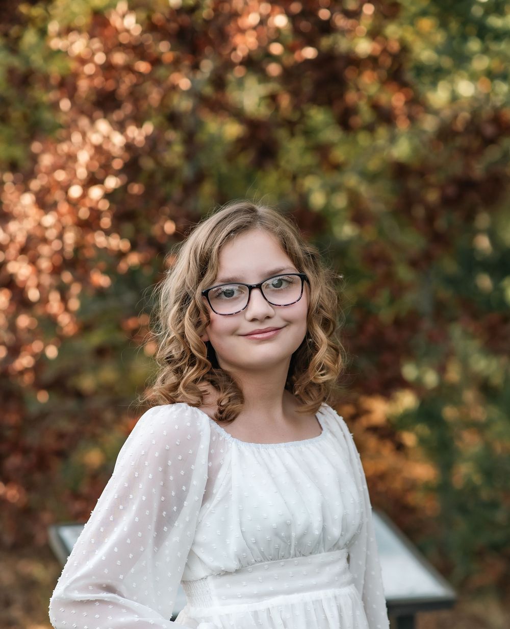 Portrait of a young person in a white dress and glasses against autumn foliage.
