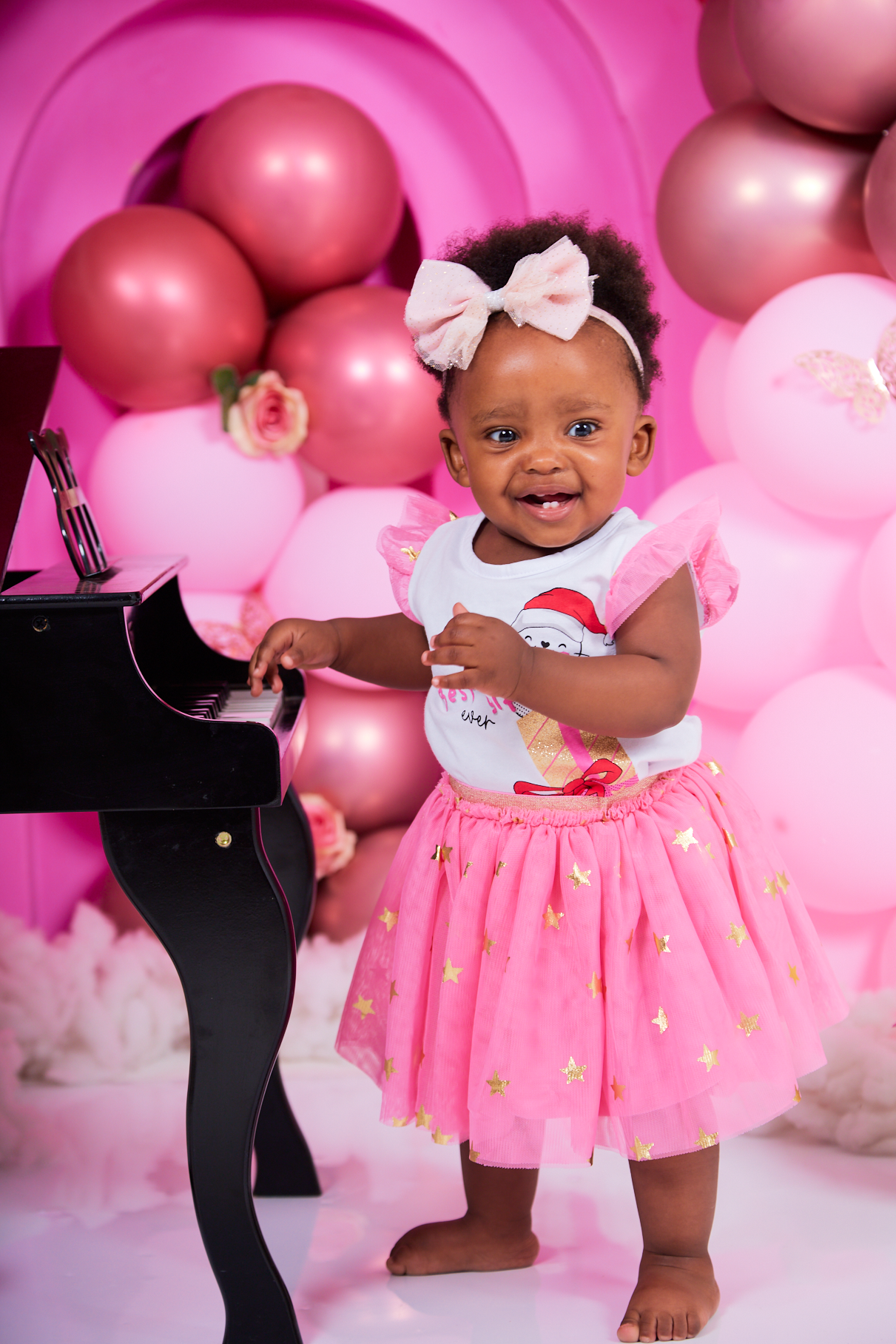 Baby in pink dress poses against pink balloon backdrop during first birthday photoshoot.