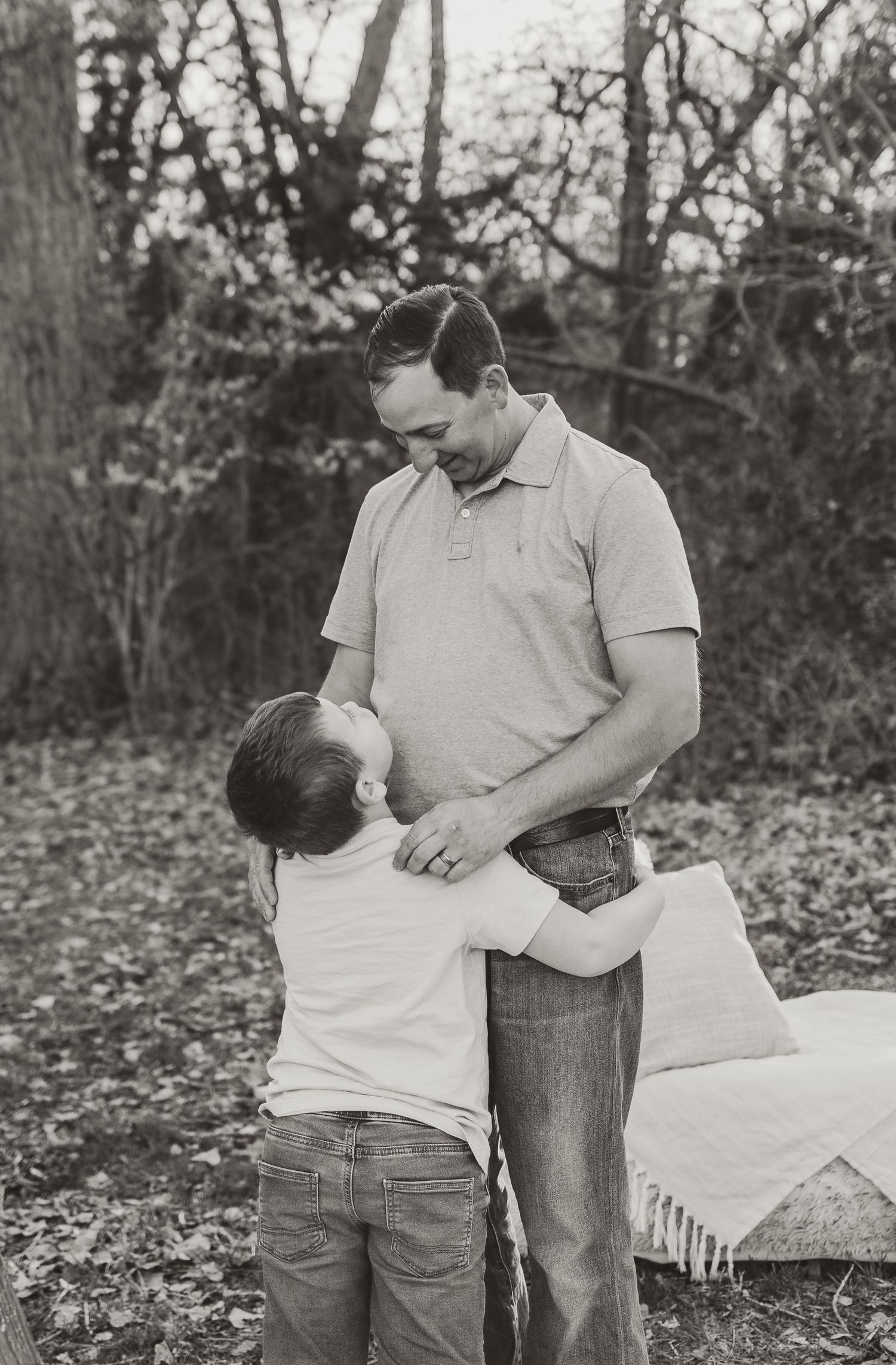 Black and white outdoor photograph showing tender moment between adult and child among trees in autumn setting.