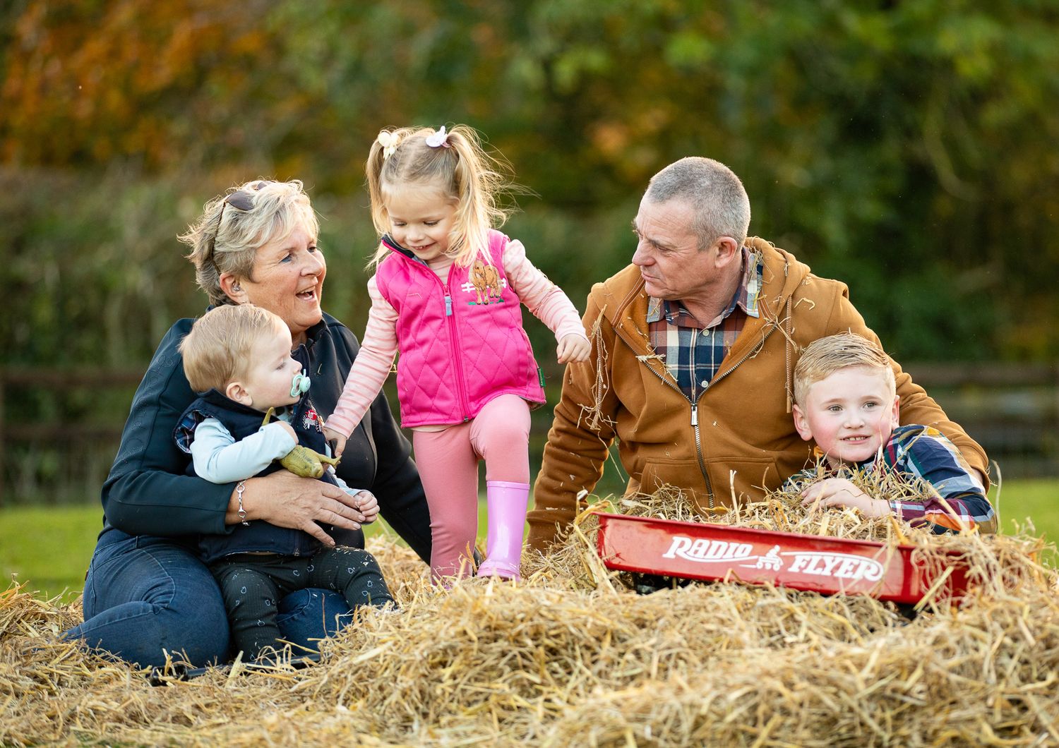 Family enjoys autumn activities together on hay bales in a garden with colorful fall foliage in background.