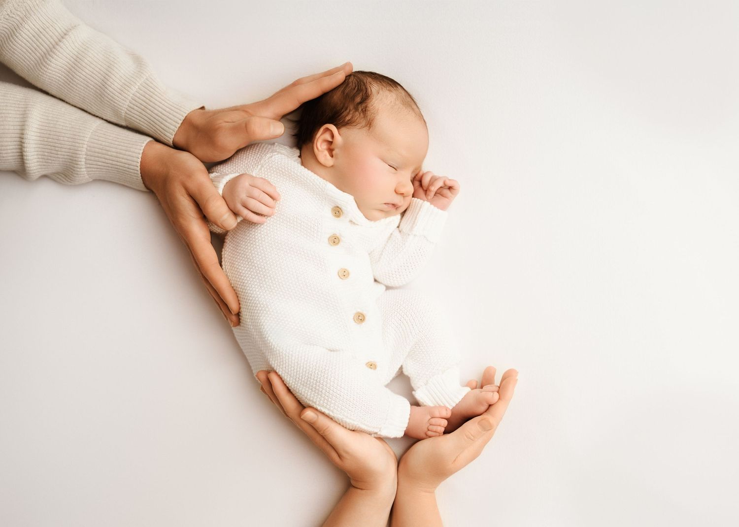 Gentle hands cradle a sleeping newborn baby wearing a white button-up onesie on a light background.