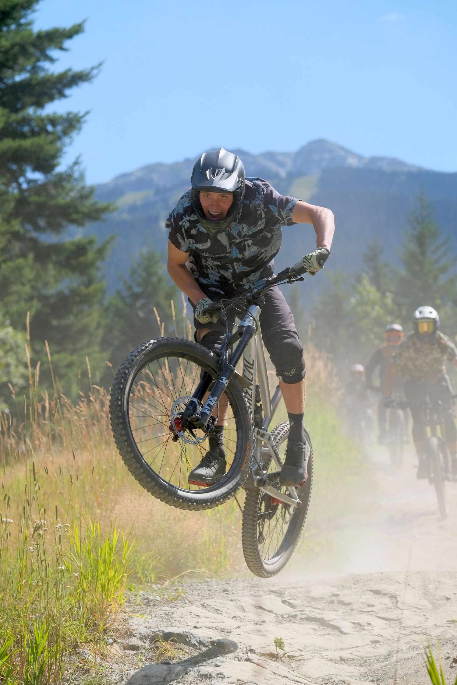 Mountain biker performing jump trick on dusty trail with mountains and evergreen trees in background.