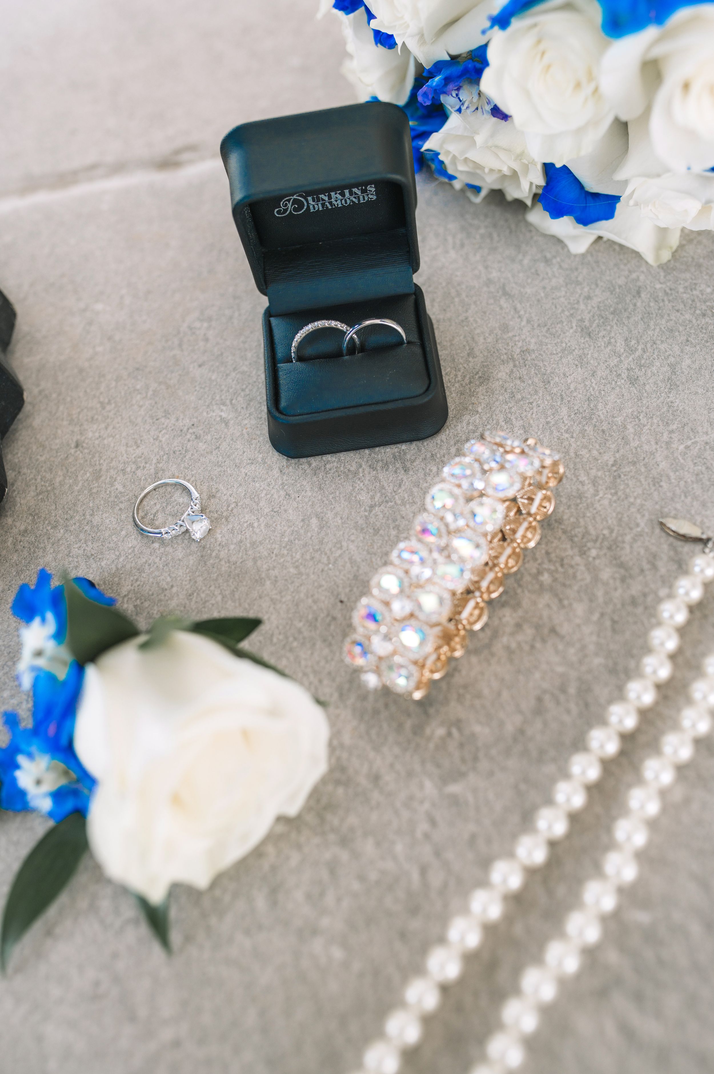 Wedding rings and jewelry displayed with white roses and pearls on gray surface.