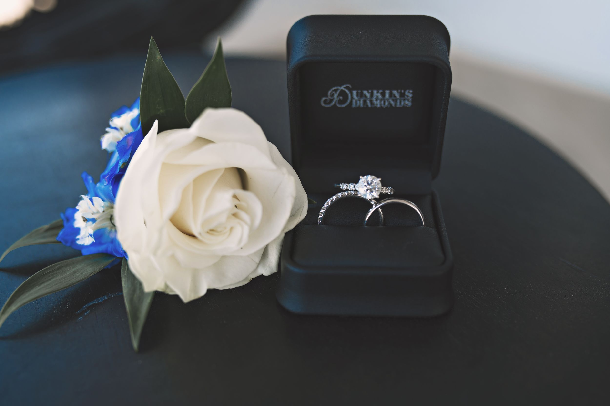 Diamond engagement ring in black box with white rose and blue accent flowers.