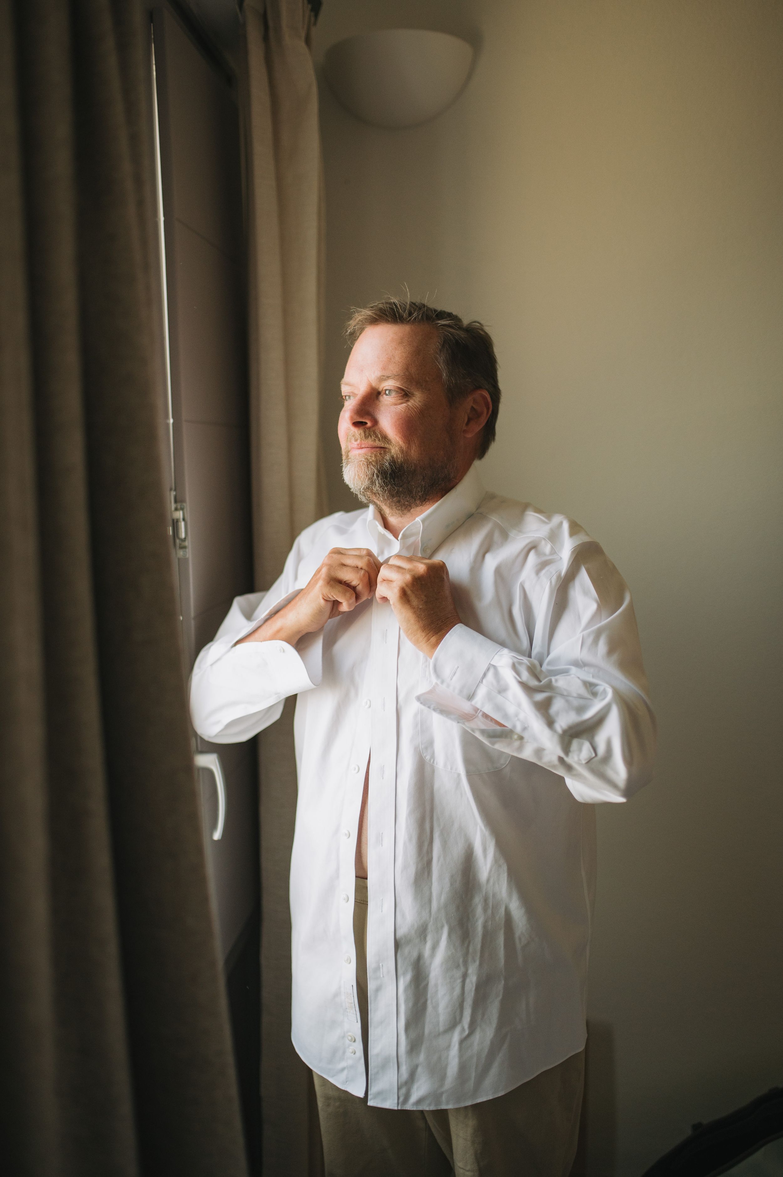 Person in white dress shirt buttoning up while getting ready near window.