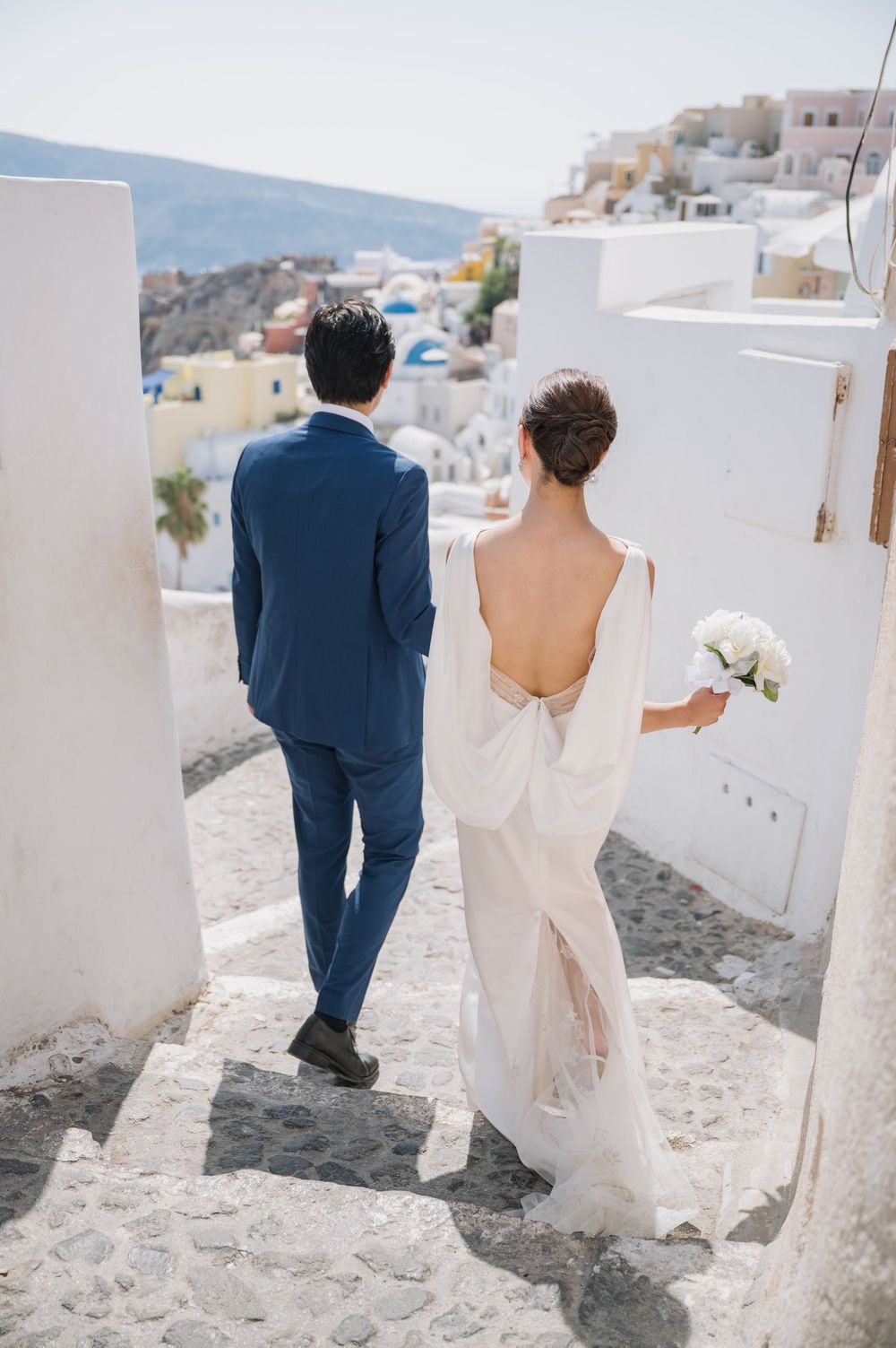 Wedding couple walks down white steps in Santorini with traditional Greek architecture in background.