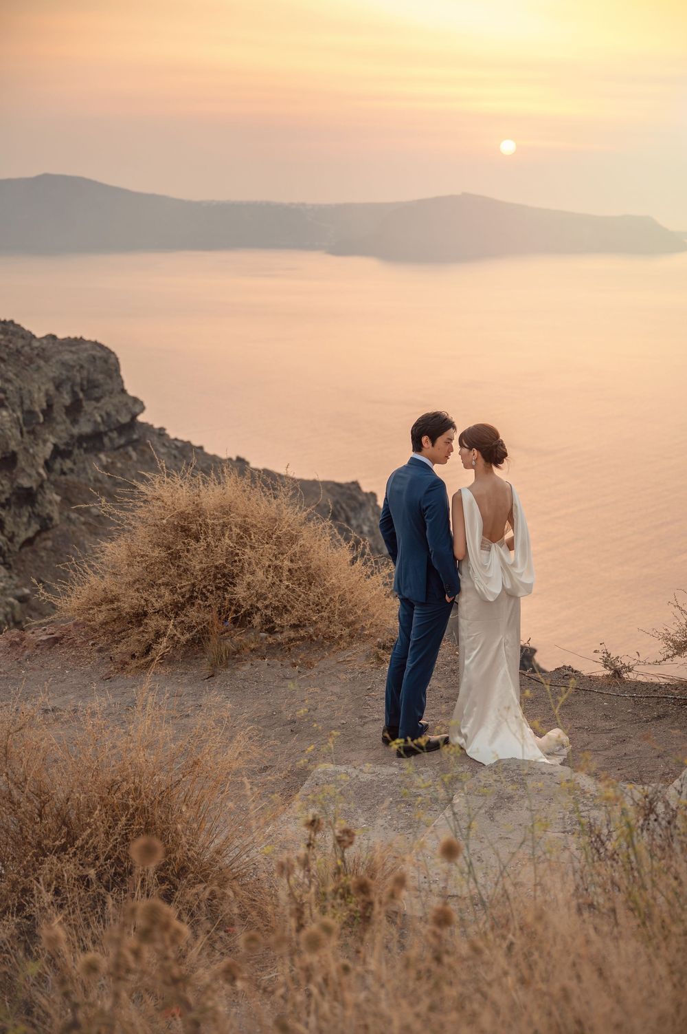 A couple stands on a cliff overlooking a misty ocean sunset.