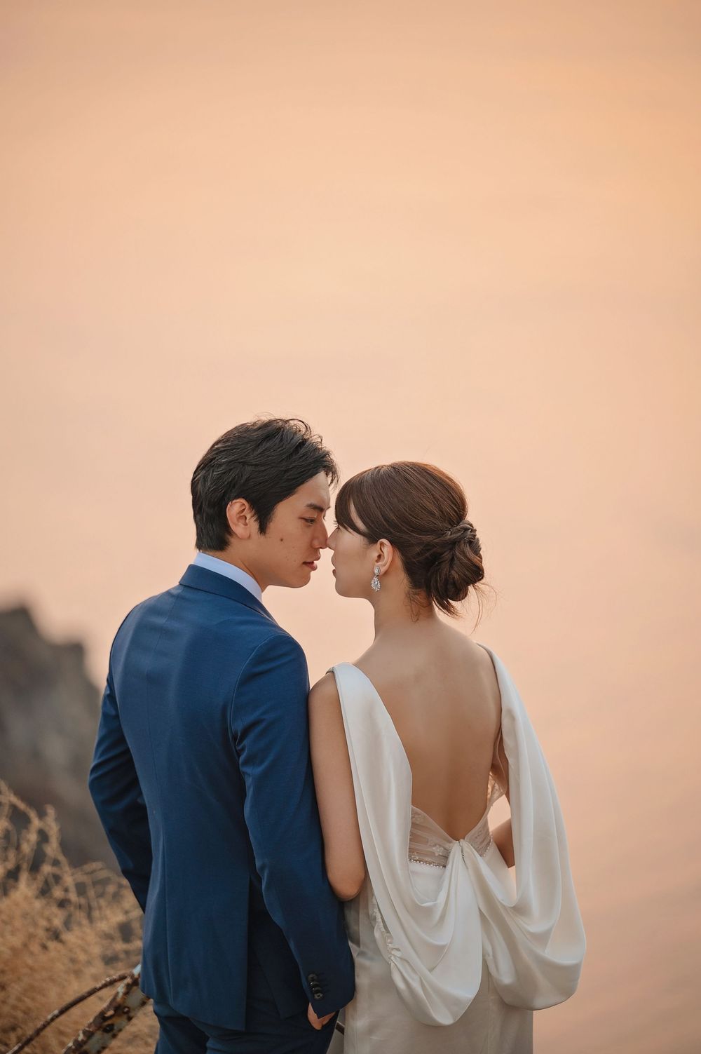 A couple embraces in elegant wedding attire during golden hour on a cliffside.