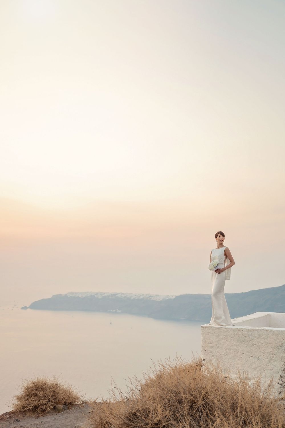 A figure in a white gown stands elegantly on a white cliffside overlooking misty mountains and sea during sunset.
