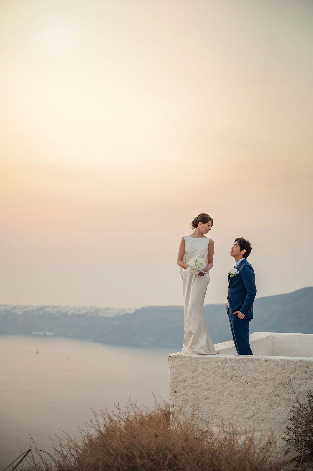 Two people stand together on a white cliffside terrace against a dreamy sunset backdrop over the Mediterranean.