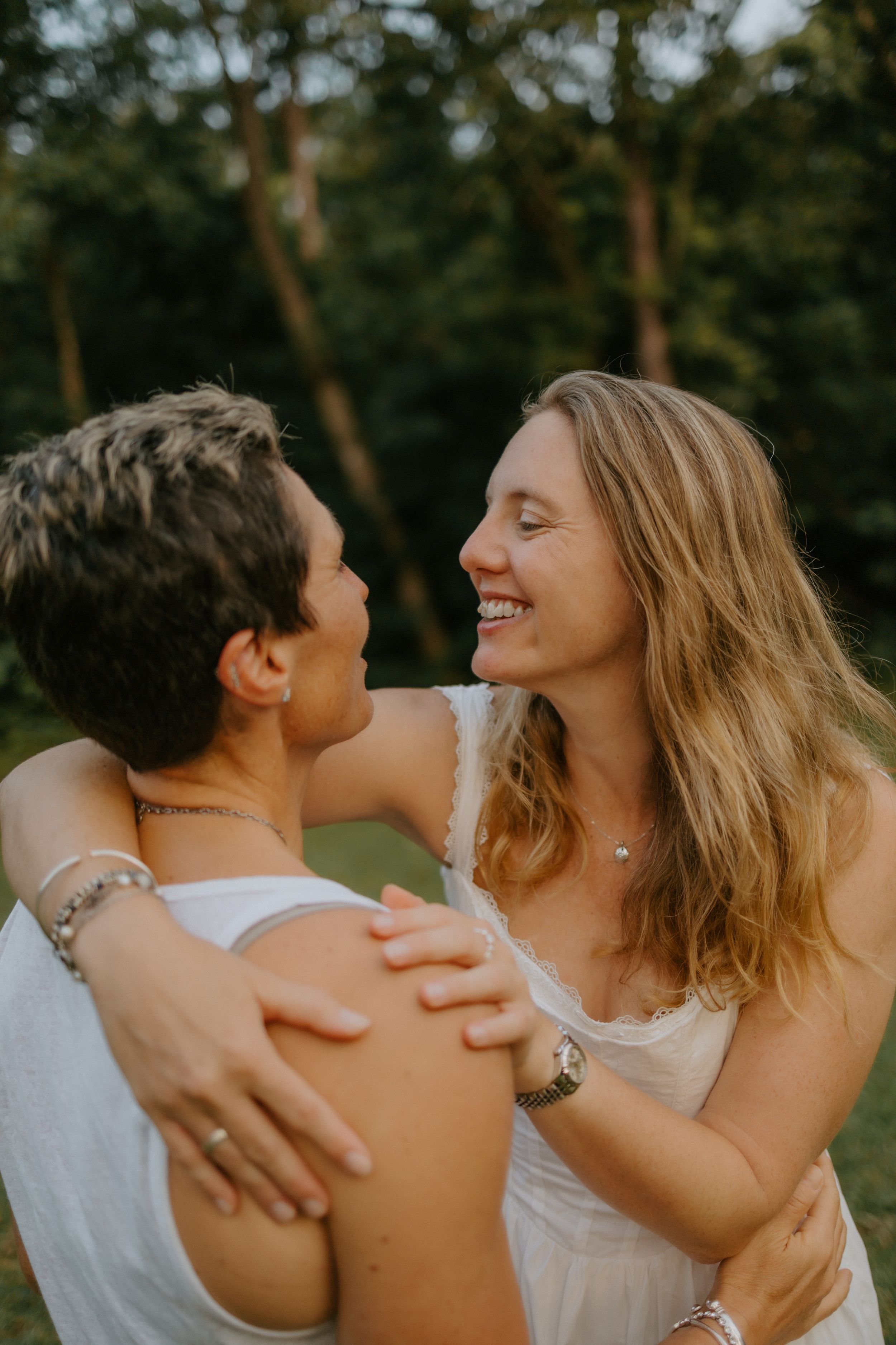 Two women embracing and smiling at each other outdoors in a lush green park setting.