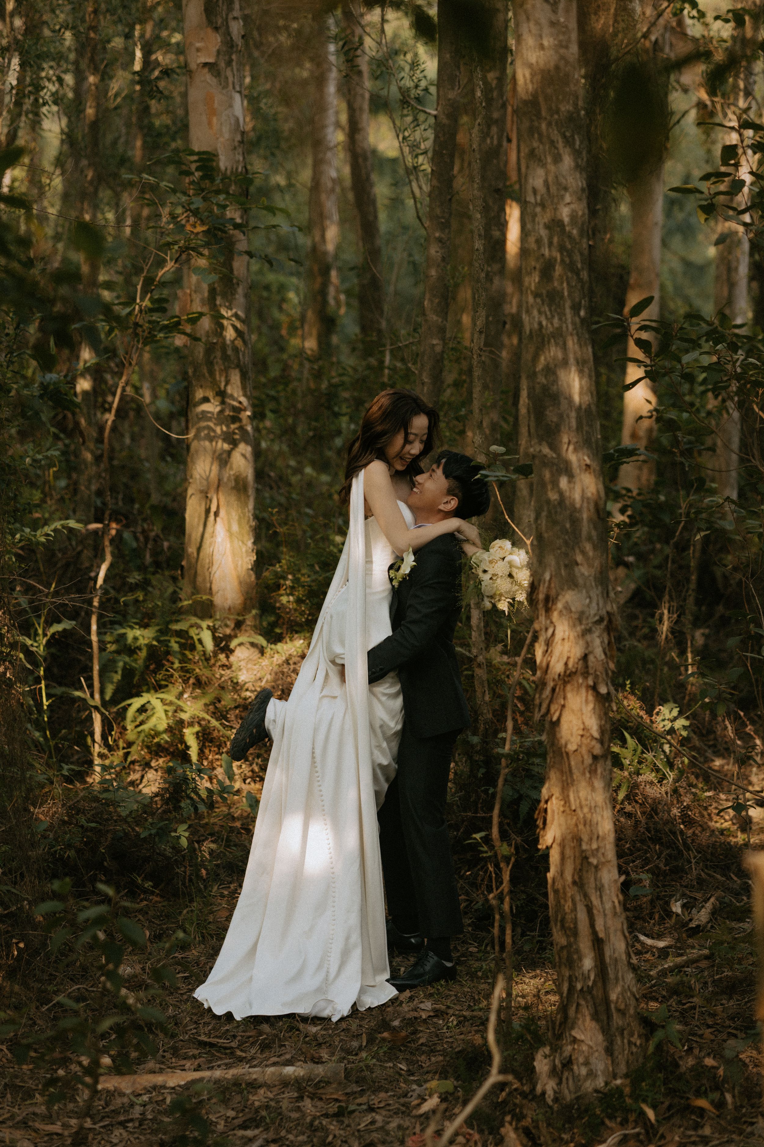 A couple embraces in a sunlit forest, the bride wearing a flowing white dress with long sleeves among tall trees.
