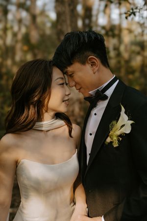 Couple in wedding attire share an intimate moment outdoors with golden sunlight filtering through autumn trees.