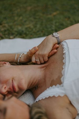 Close-up of couple holding hands while lying on white blanket outdoors, woman wearing silver watch and rings.