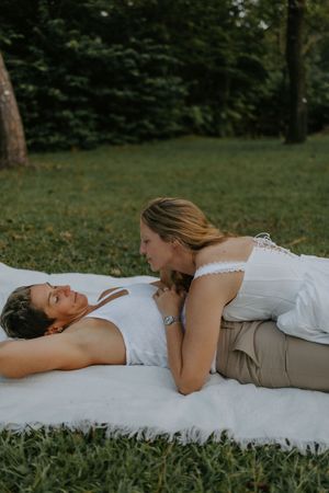 Two women share a romantic moment lying on a white blanket in a lush green park setting.