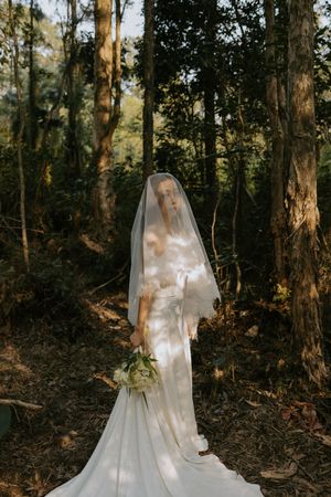 A bride in an elegant white wedding dress and long veil stands in a sunlit forest holding a bouquet.
