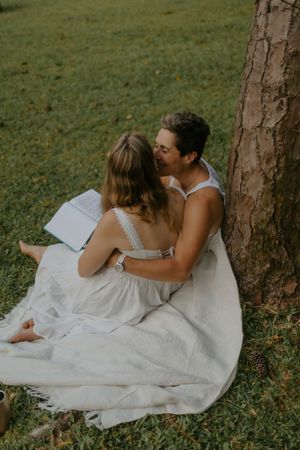 Couple sitting together against a tree on a white blanket in a park, sharing a tender kiss while reading a book.