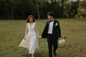A couple in wedding attire walks hand in hand through a grassy field during golden hour at sunset.