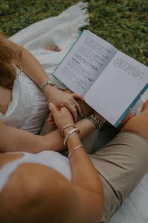 Couple holding hands while reading a journal together on a blanket outdoors in a park.