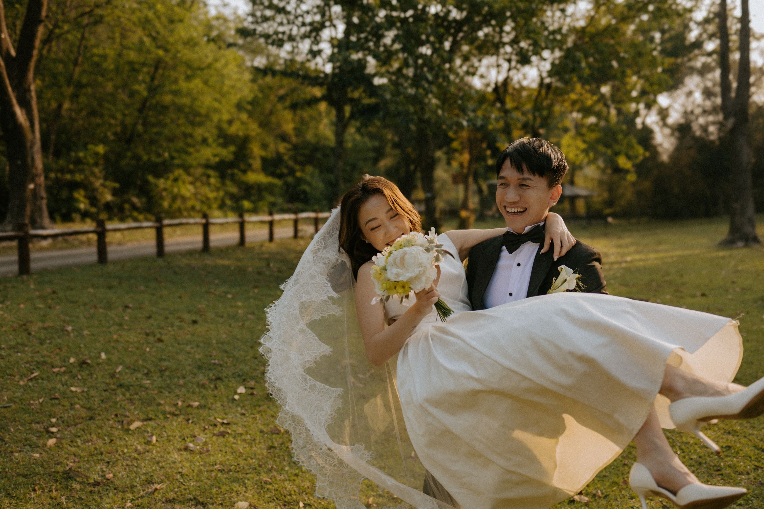 A romantic wedding photo captured in a lush park setting during golden hour with floating white dress and bouquet.