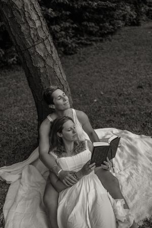 Two women in white dresses sit against a tree in a black and white photo, one reading a book outdoors.