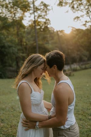 Couple sharing a romantic forehead touch embrace at golden hour sunset in a lush green park setting.