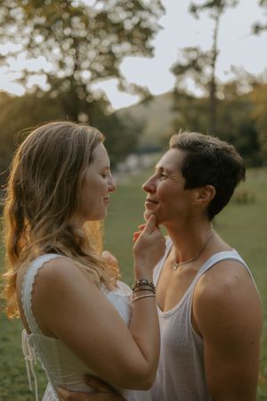Two women share a tender moment outdoors at golden hour, one gently touching the other's chin.