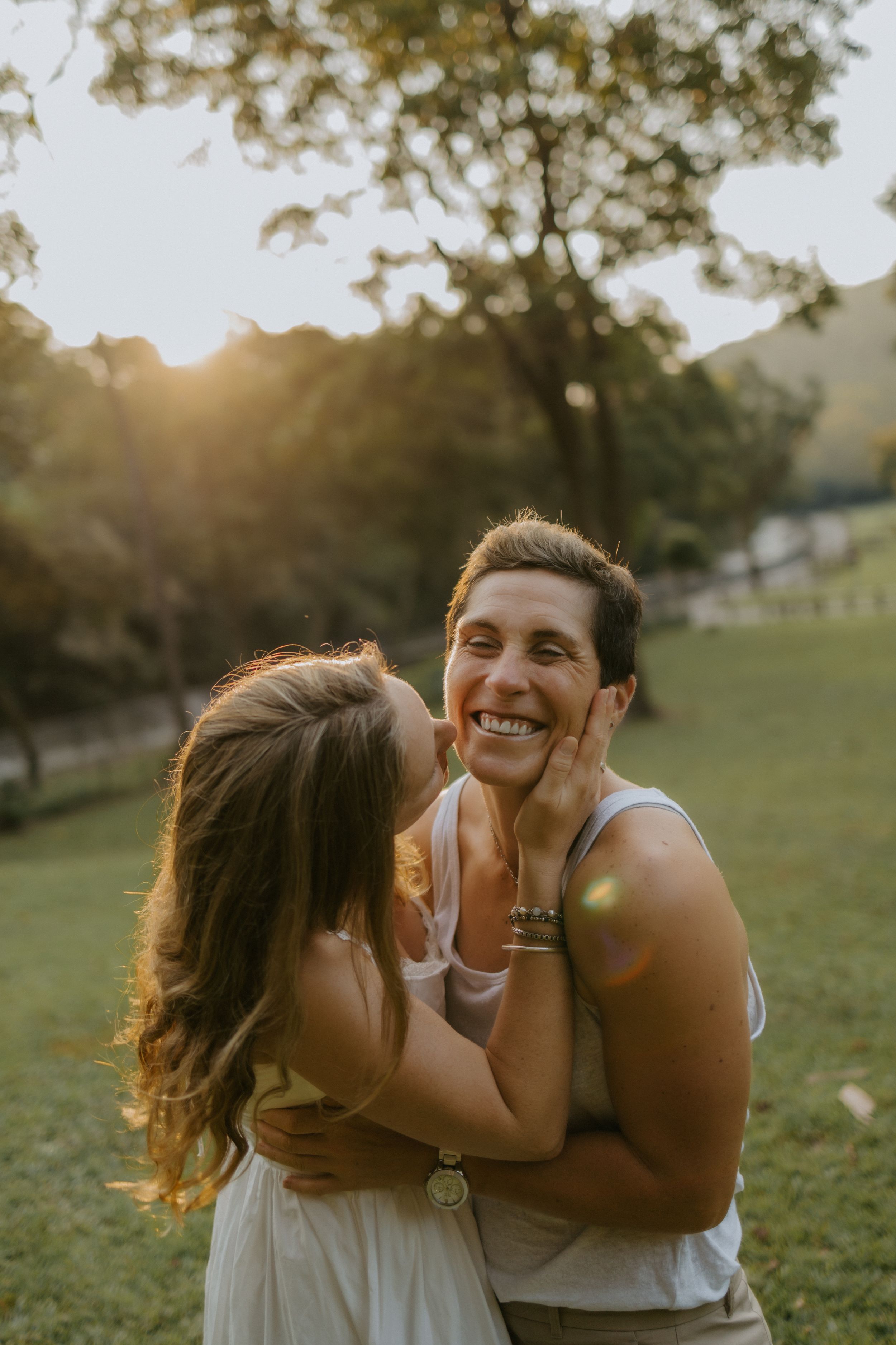 Two women share a joyful embrace outdoors at golden hour, smiling warmly in a sunlit park setting.