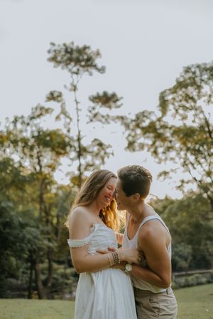 A couple shares a romantic moment outdoors at golden hour, surrounded by lush green trees in a park setting.