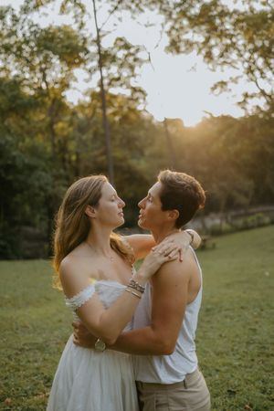 Two women embrace and gaze at each other during a golden sunset in a lush green park setting.