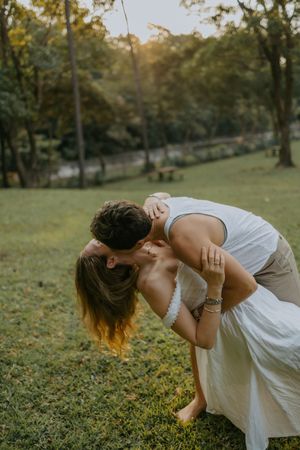 Couple sharing a romantic dip kiss in a lush green park at golden hour, woman in white dress.