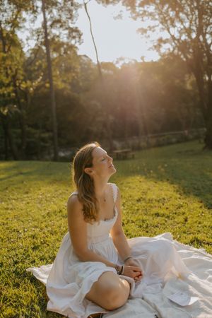 Young woman in white dress sitting on grass blanket, basking in golden sunset light in a lush green park.