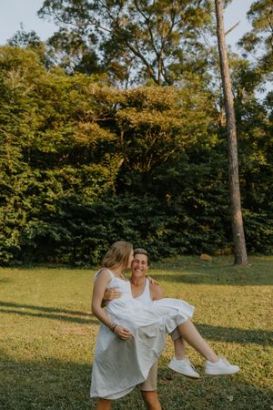 Two women in white dresses share a joyful moment outdoors, one carrying the other in a lush green park setting.