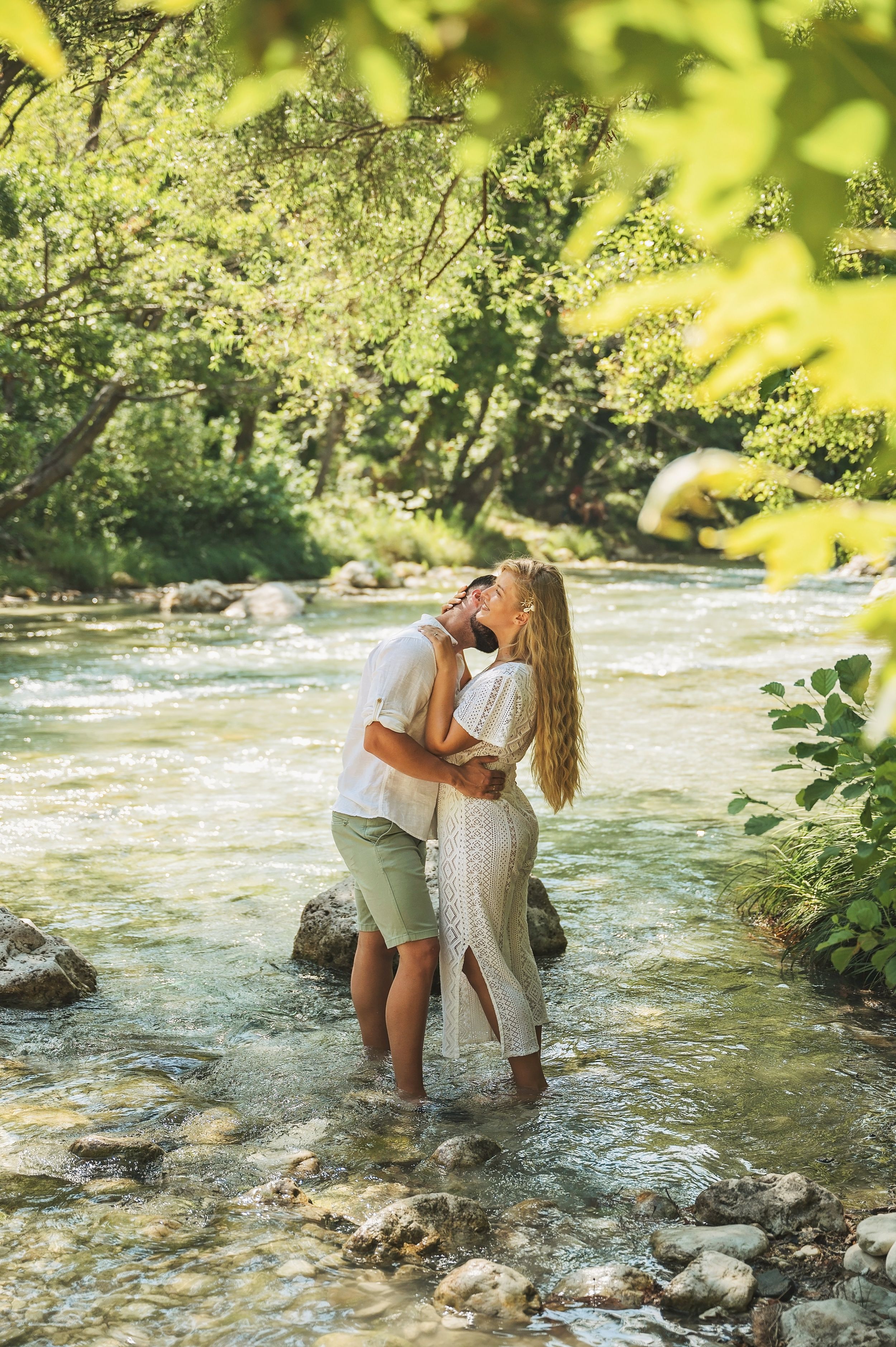 A couple shares a kiss while standing in a sunlit river with green foliage overhead.