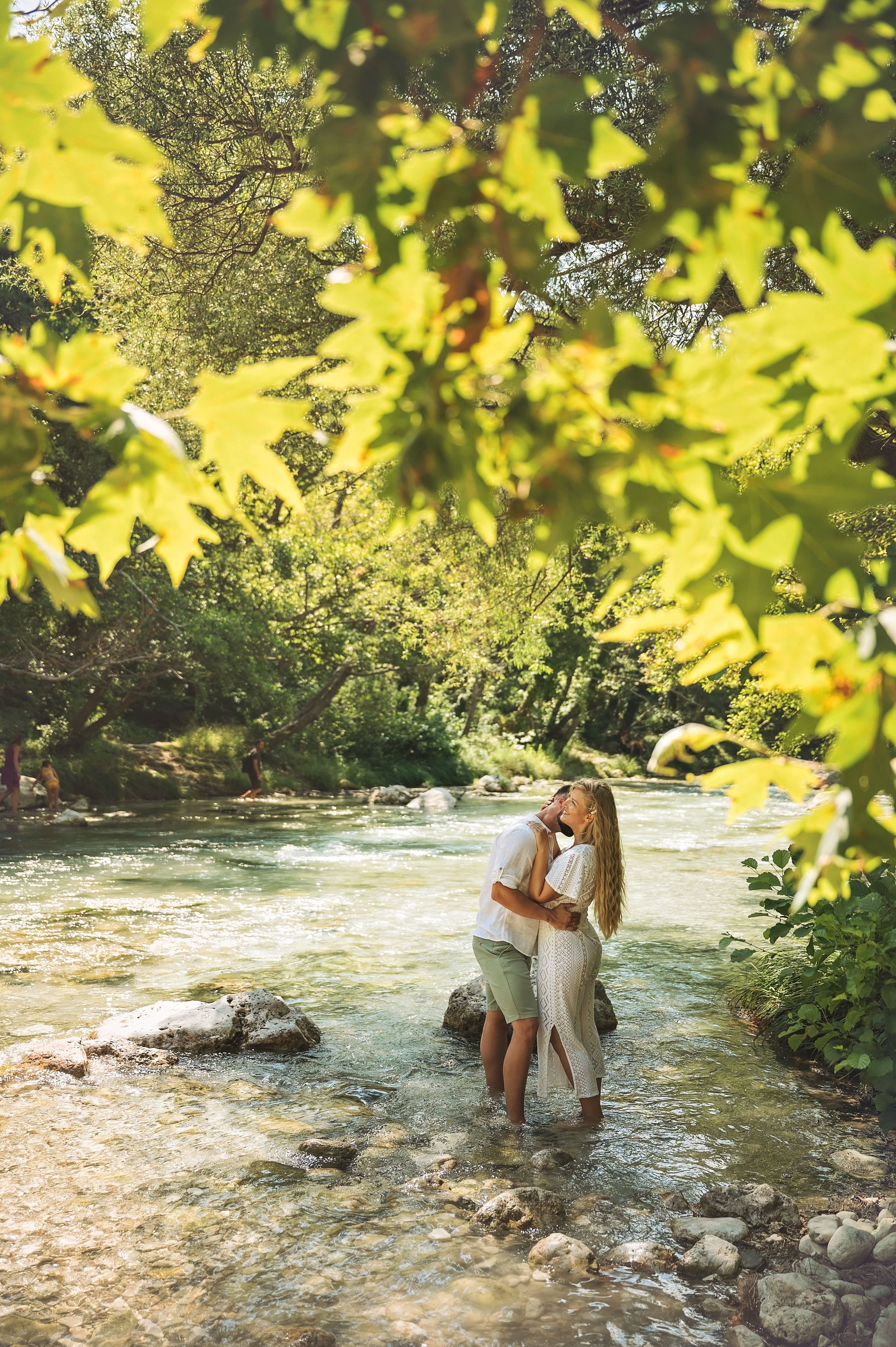A couple embraces while standing in a shallow river surrounded by yellow autumn foliage and sunlight.