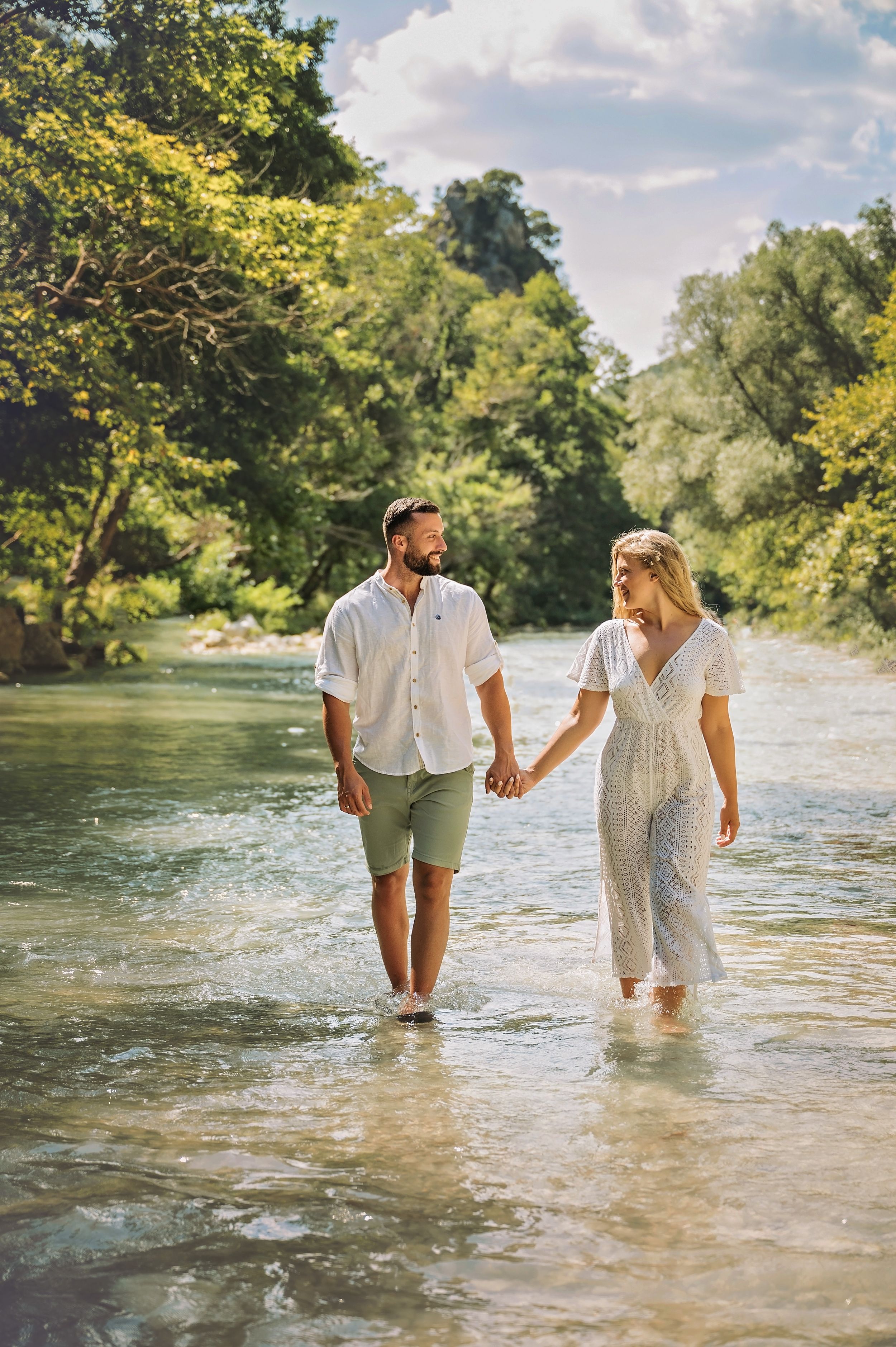 A couple holds hands while walking through crystal clear river water surrounded by lush greenery.