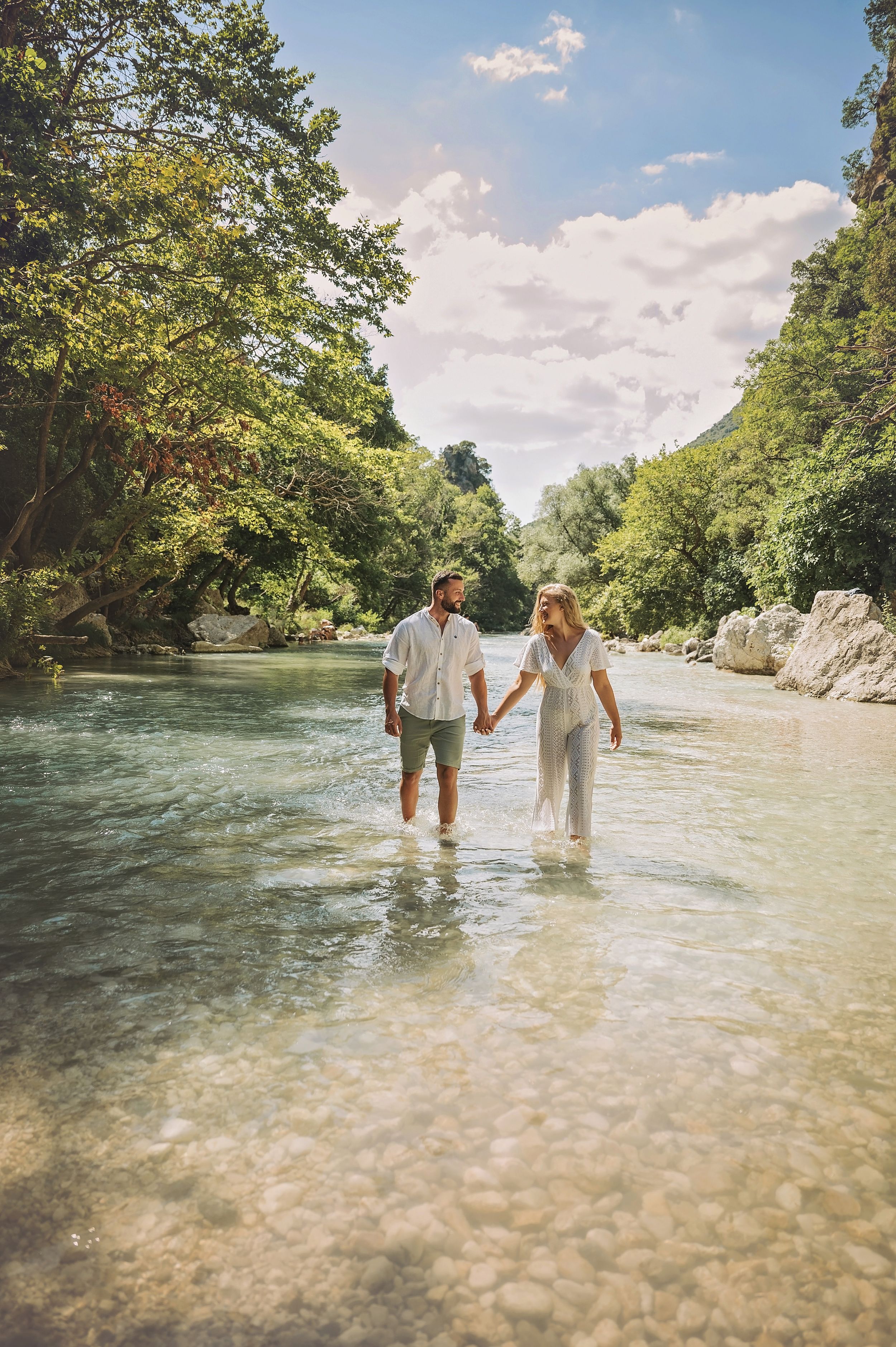 Couple wading together in a scenic river surrounded by trees under a bright blue sky.