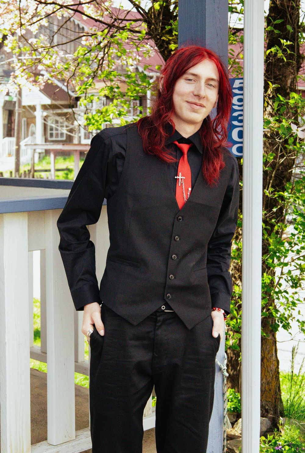 Person in formal black vest and tie poses against outdoor backdrop with blooming spring flowers.