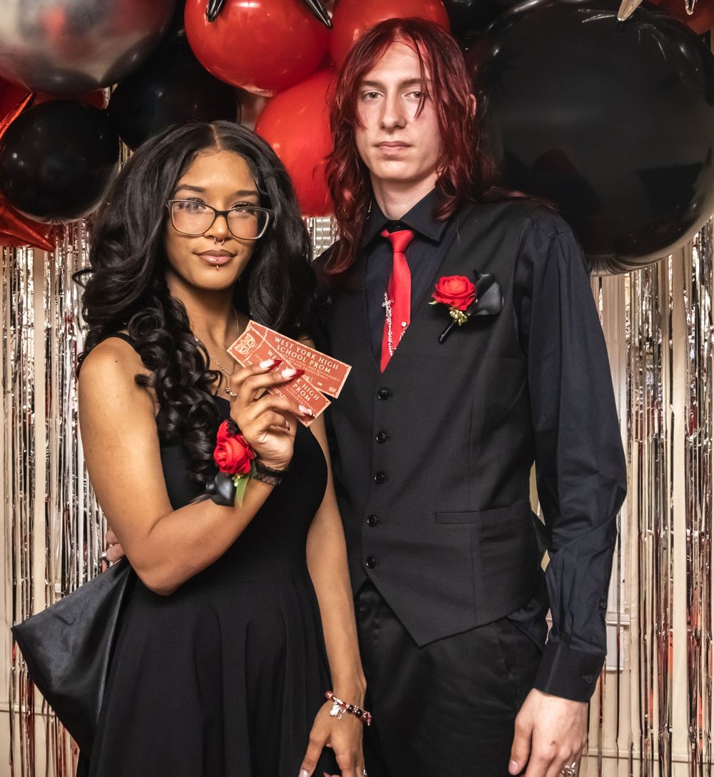 Two people in formal black attire pose together with red balloons and silver tinsel backdrop for special occasion.