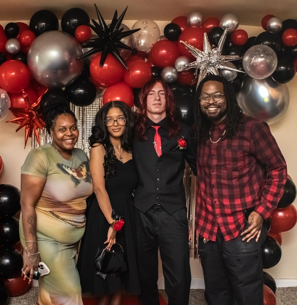 A group poses together at a party with red, black, and silver balloon decorations in the background.
