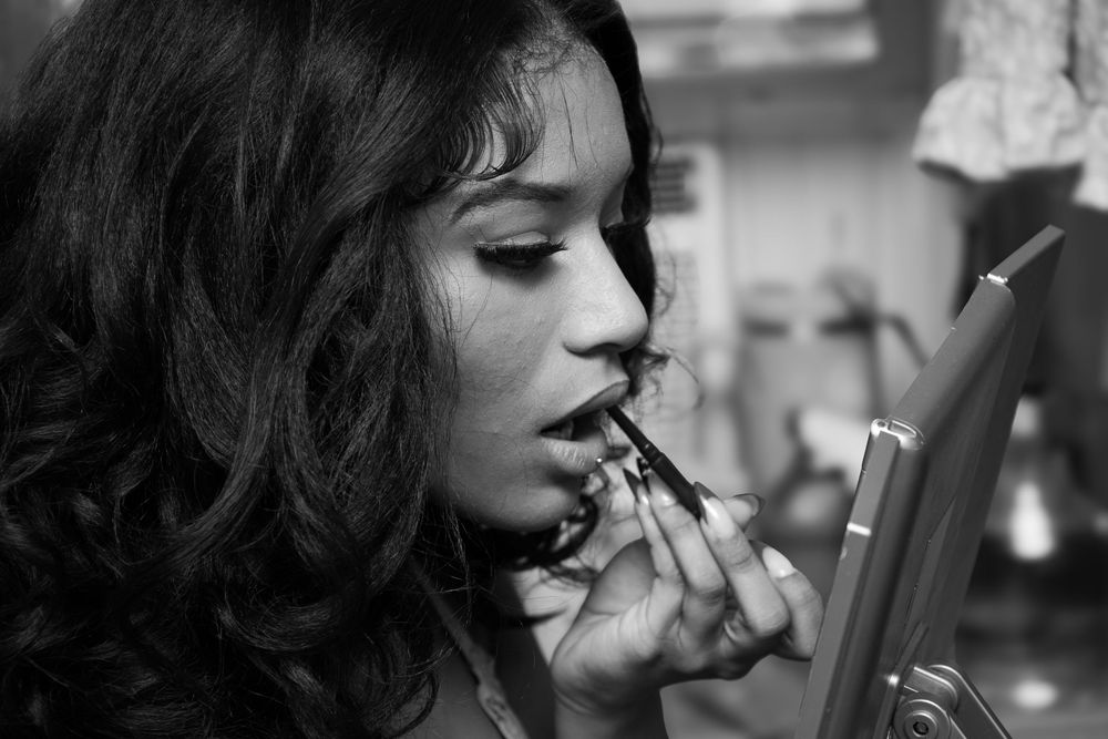 A woman in black and white applies lipstick while looking in a mirror, with dark curls framing her face.