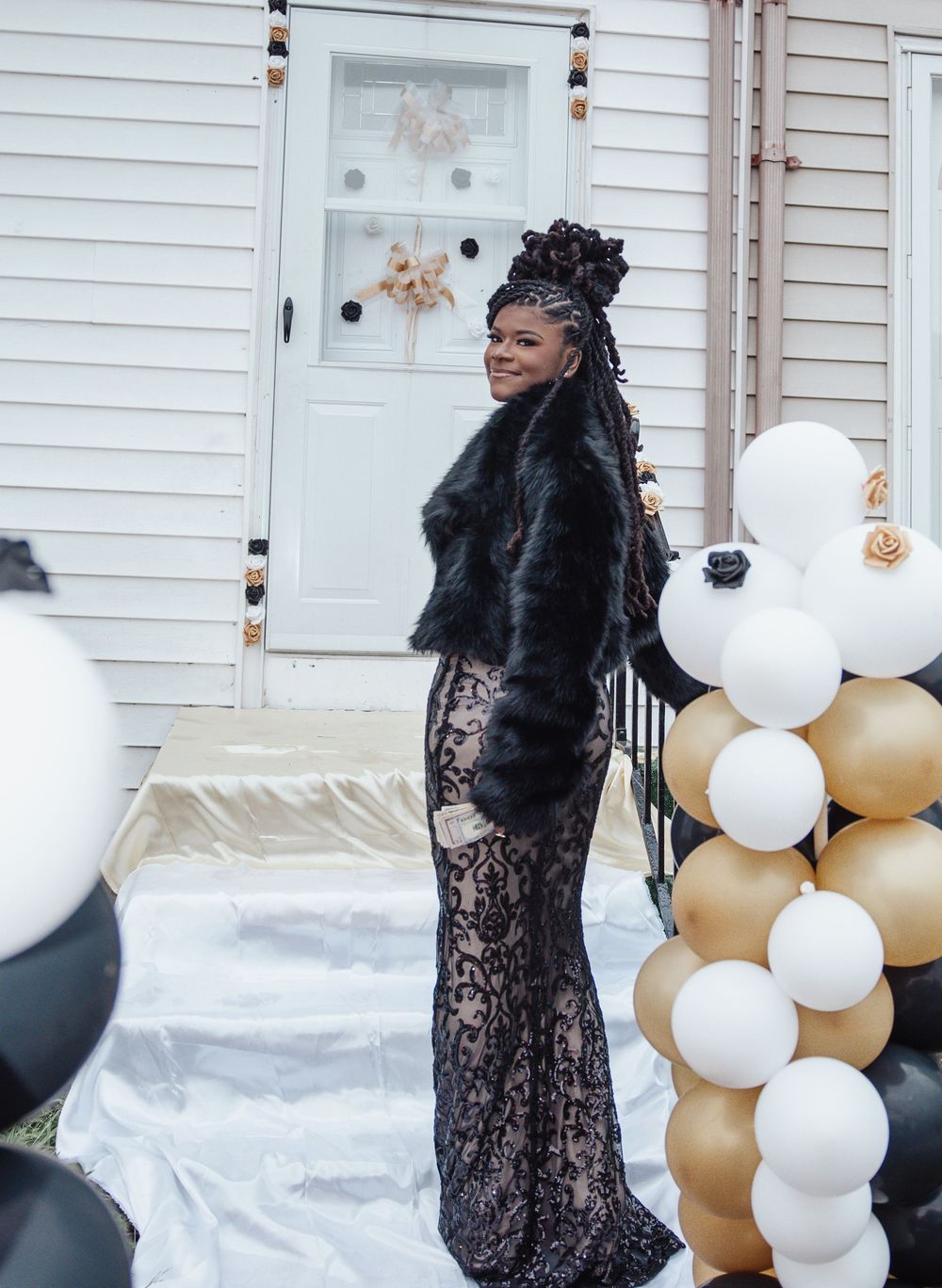 Formal dress event setup with black and gold balloon arch and white siding house backdrop.