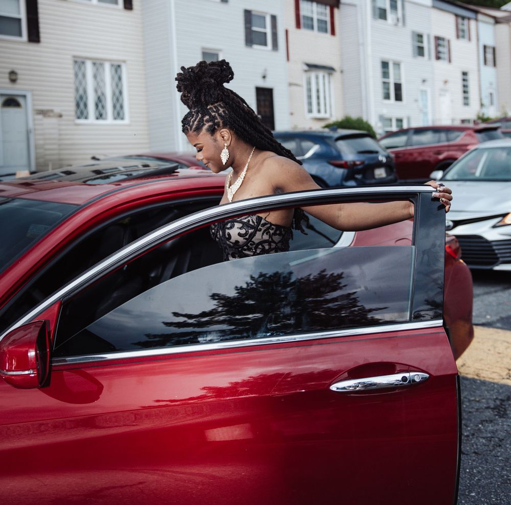 Someone in an elegant dress emerging from a red car door in an apartment complex parking area.