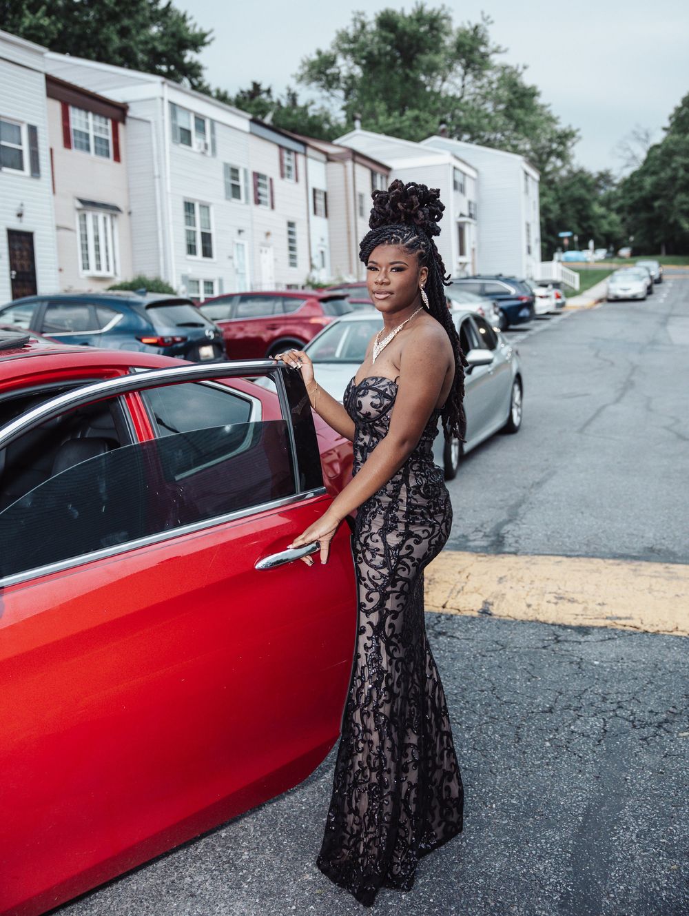 Person in elegant black lace gown poses next to red car on residential street with townhouses in background.
